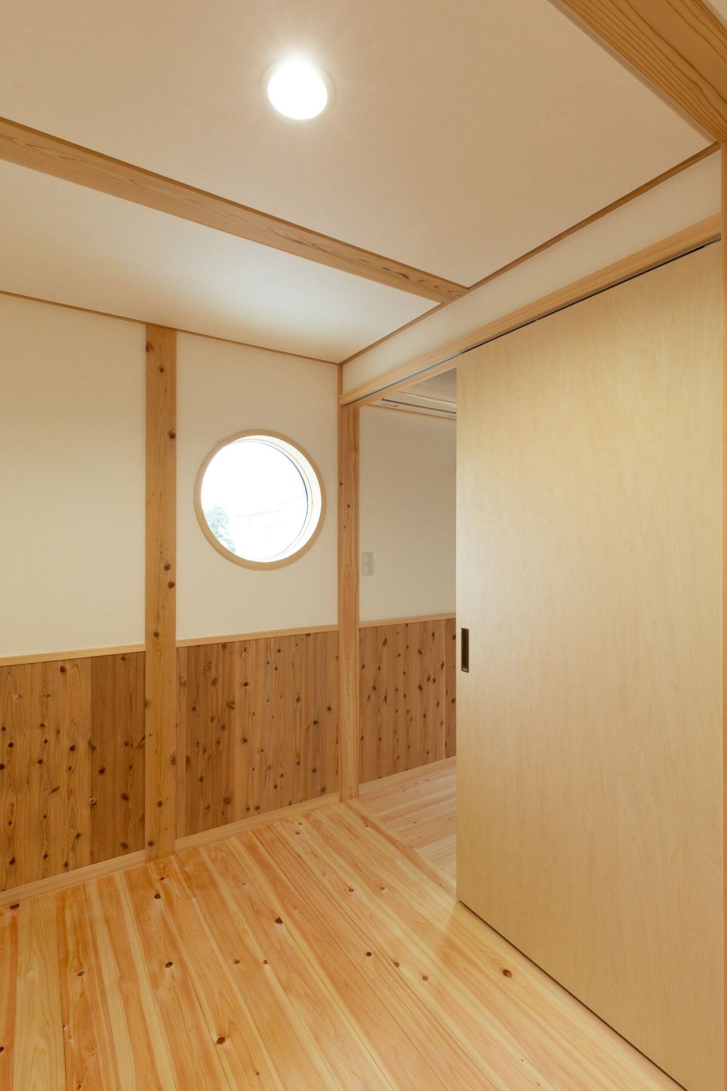 無垢材の床と腰壁、丸窓から光が差し込む廊下と木製引き戸の部屋の入口